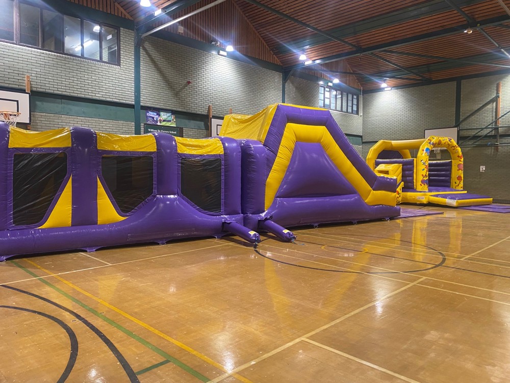 Large purple and yellow inflatable obstacle course and matching bouncy castle set up in sports hall