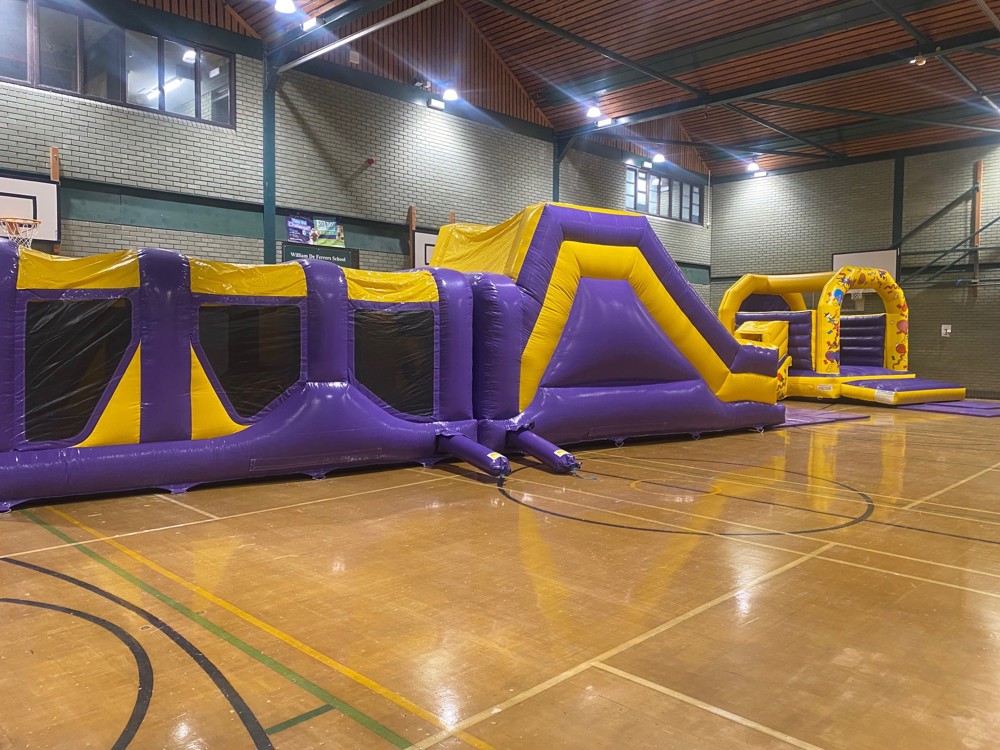 Large purple and yellow inflatable obstacle course and matching bouncy castle set up in sports hall