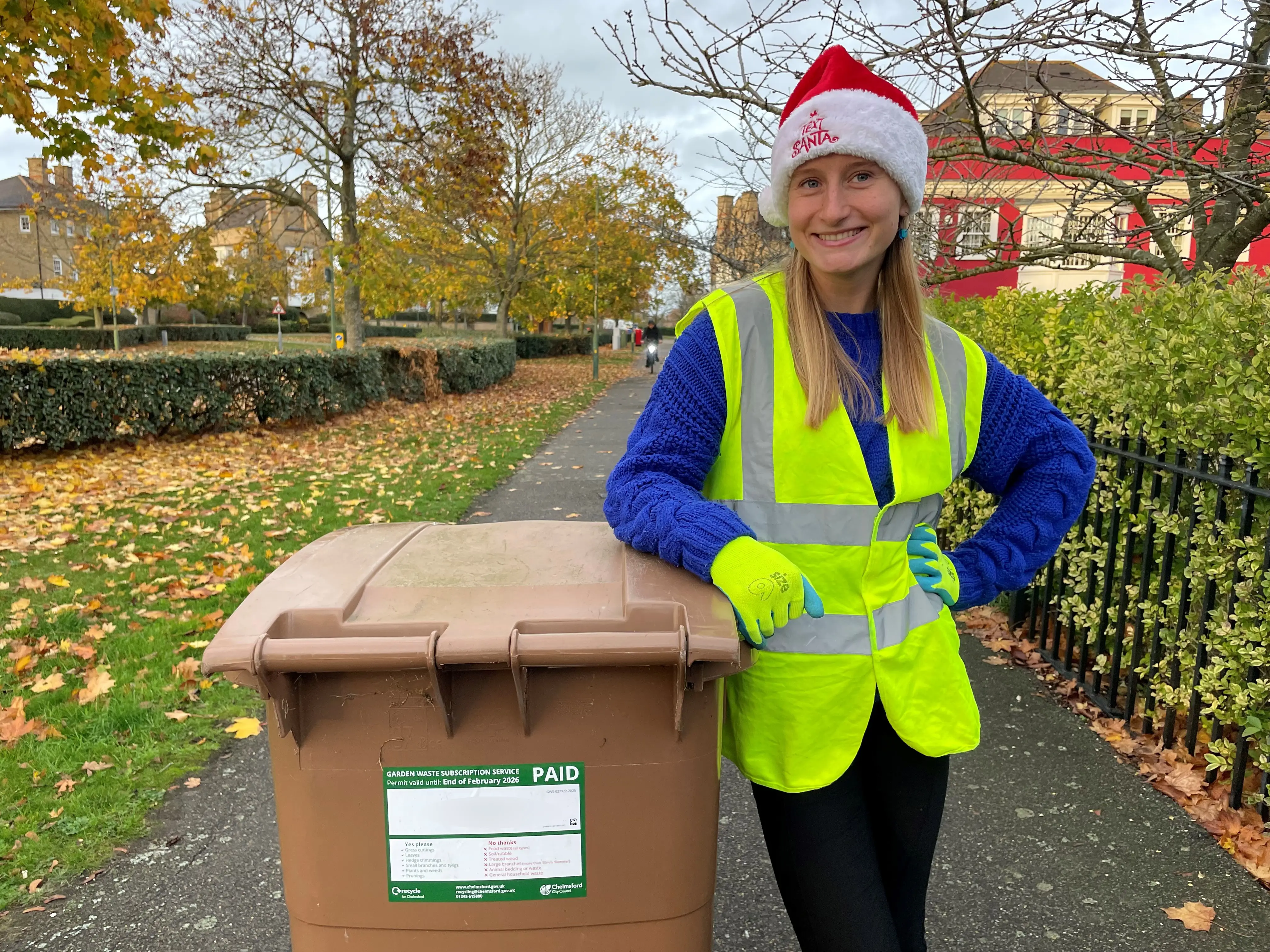 Woman in yellow high-vis jacket wearing Santa hat and standing with brown wheelie bin