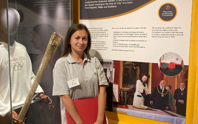 A woman with long, dark hair smiling in front of a London Olympics 2012 uniform on display. 