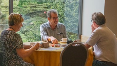 Two men and a woman sit and talk around a round table while having a cup of tea or coffee. 