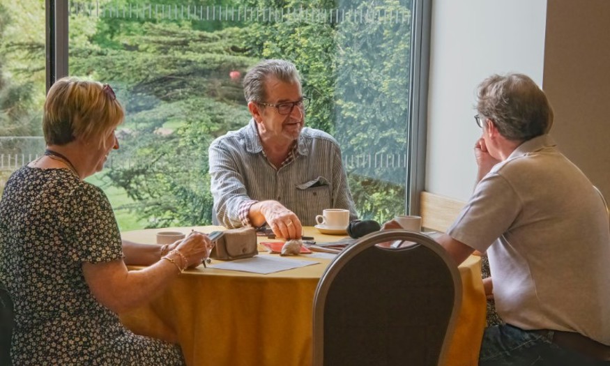 Two men and a woman sit and talk around a round table while having a cup of tea or coffee. 