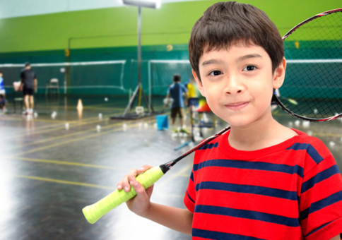Young boy holding badminton racket in front of courts with other people playing