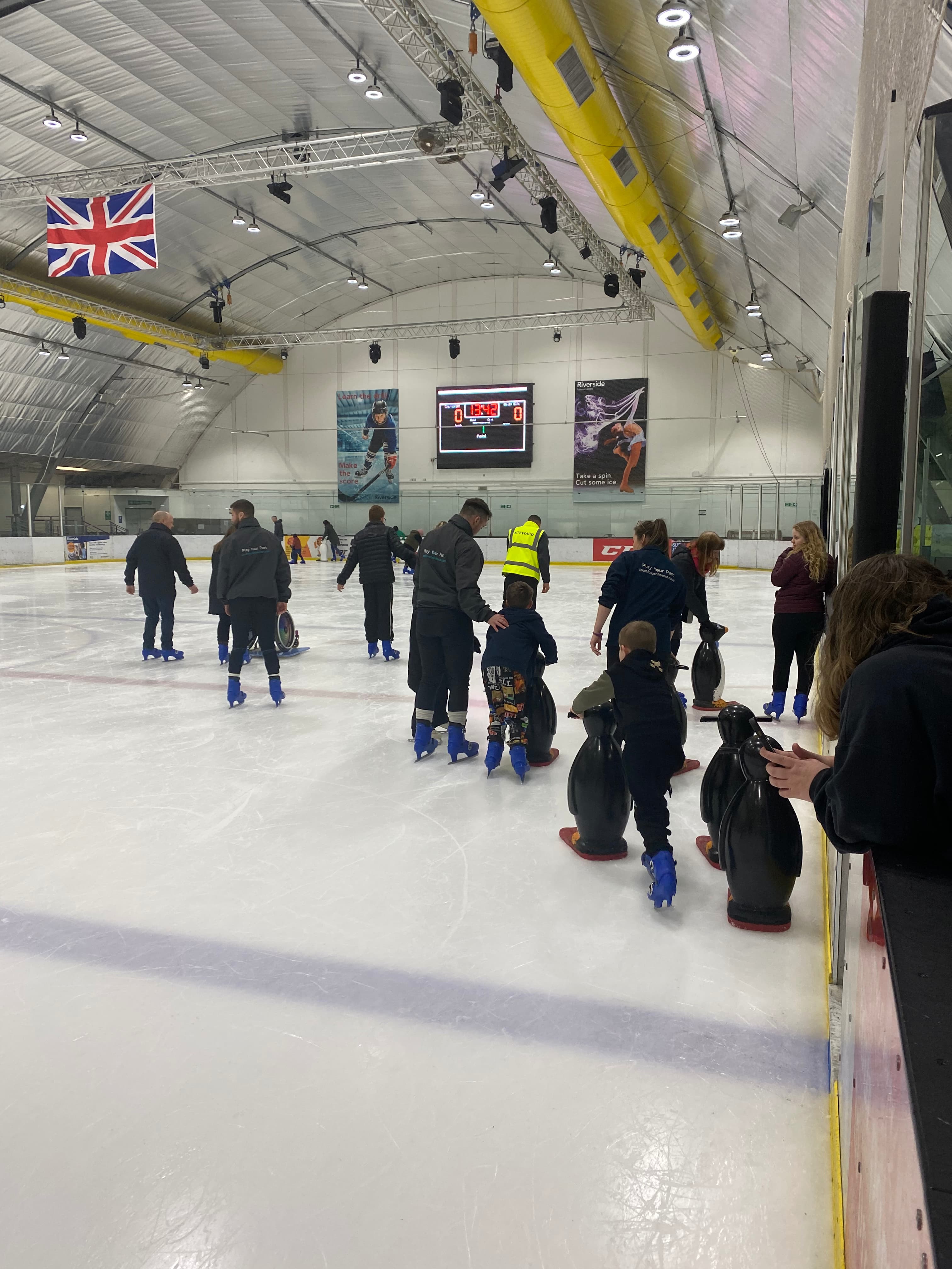 Parents and children ice skating with scoreboard on the wall in the background