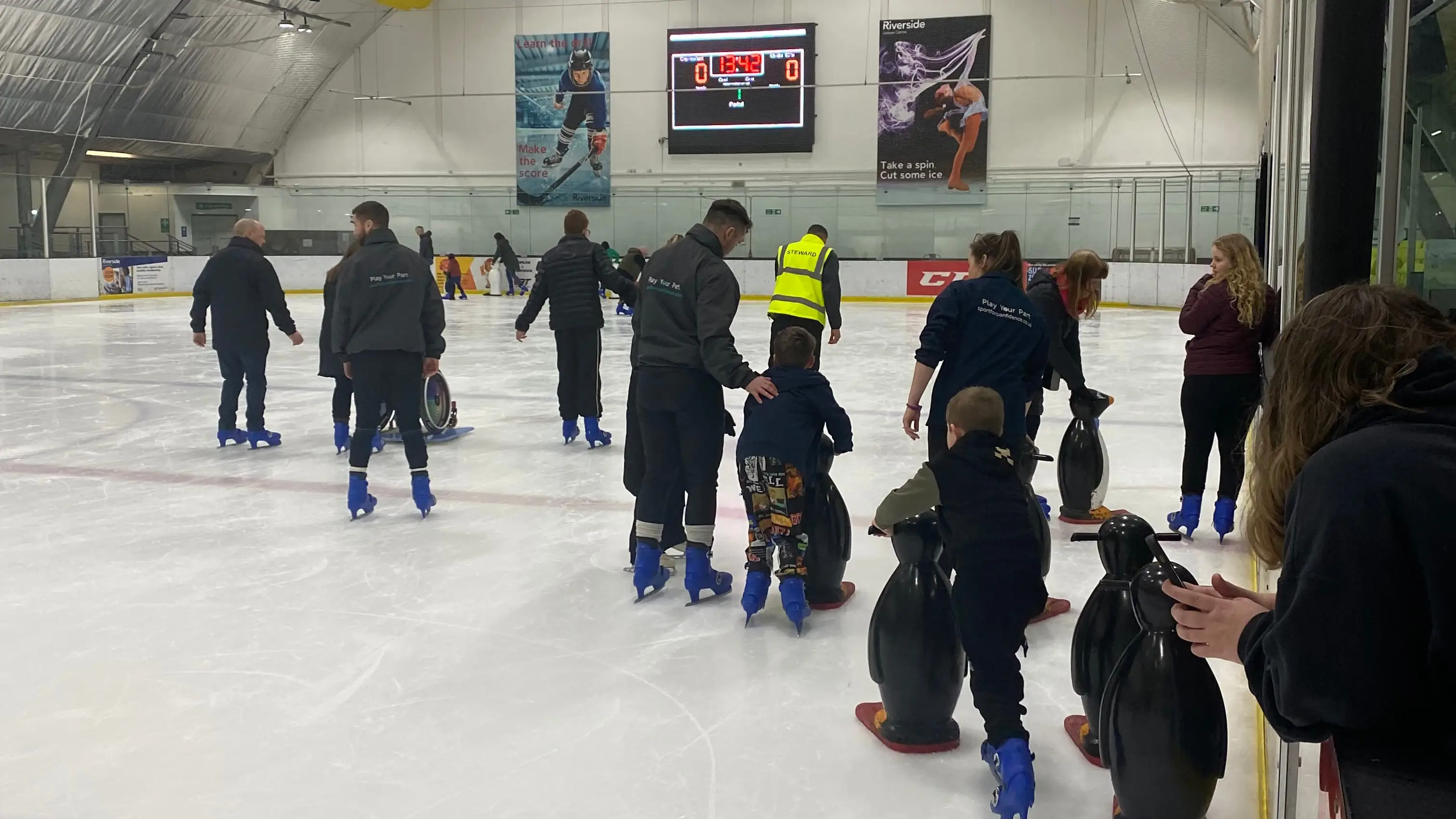 Parents and children ice skating with scoreboard on the wall in the background
