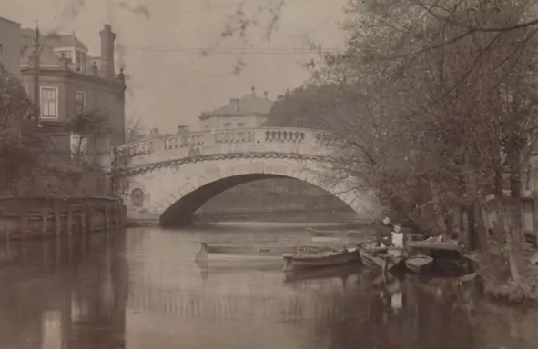 Small wooden boats on river in front of bridge with large single arch (old black and white photo)