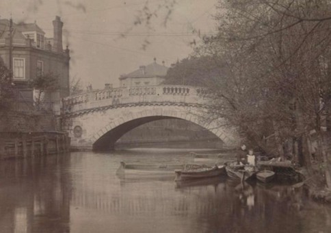 Small wooden boats on river in front of bridge with large single arch (old black and white photo)
