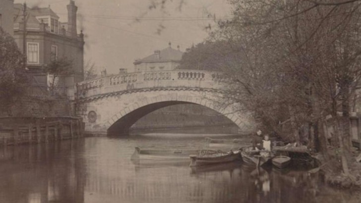 Small wooden boats on river in front of bridge with large single arch (old black and white photo)