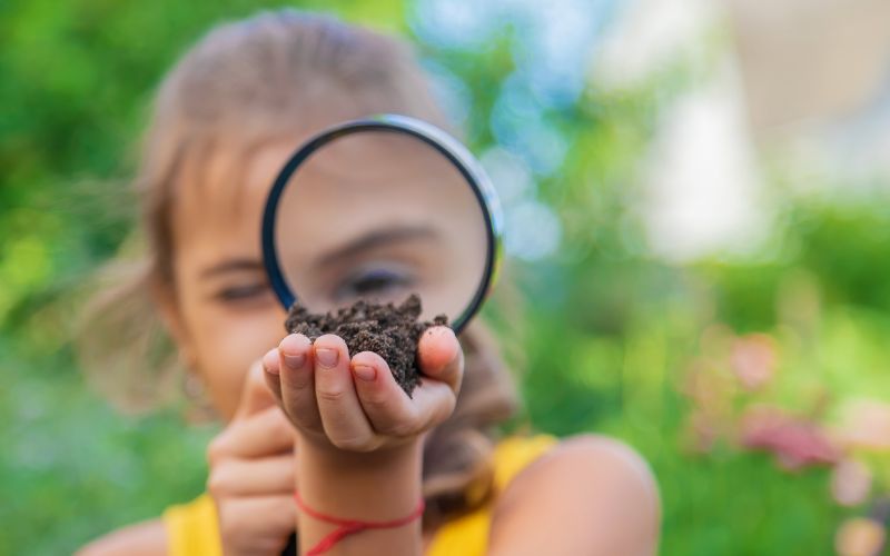 A young girl holds up a magnifying glass to her eye and looks at soil that she is holding in her hand. 