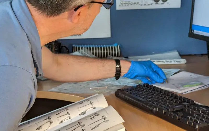A man sat at a desk in front of a computer. He is inspecting an item while referencing a book. 